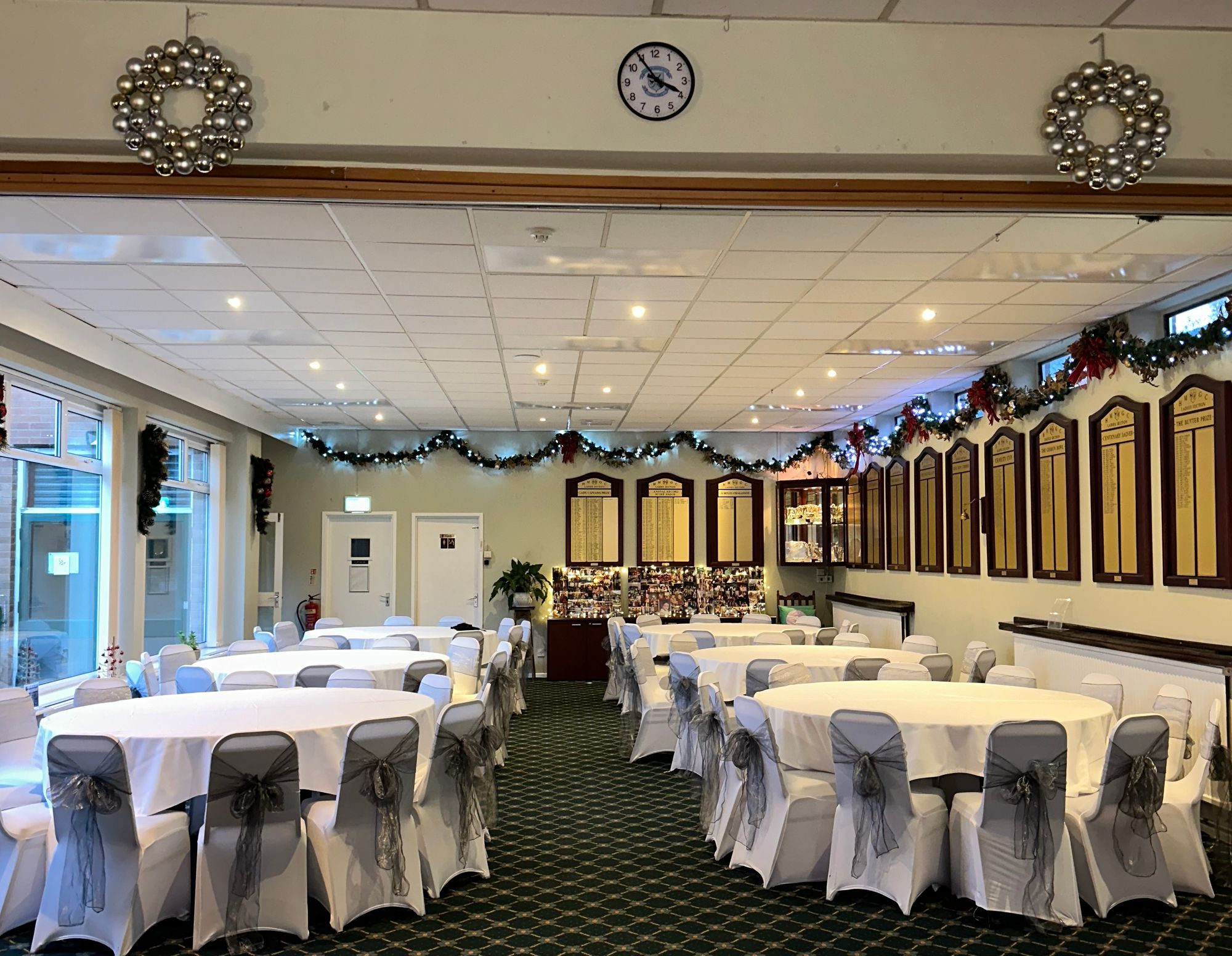 Event hall with round tables covered in white cloths, chairs with gray bows, decorated with Christmas garlands and wreaths.