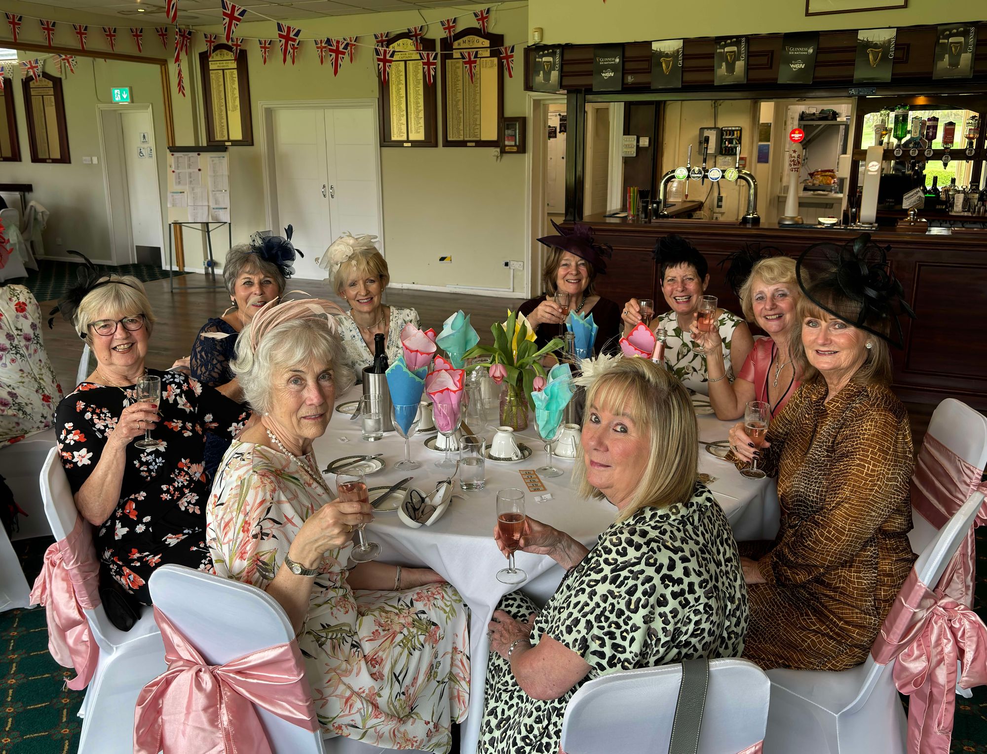 Group of elderly women dressed elegantly with hats sitting around a table holding glasses of rosé wine in a decorated indoor setting.
