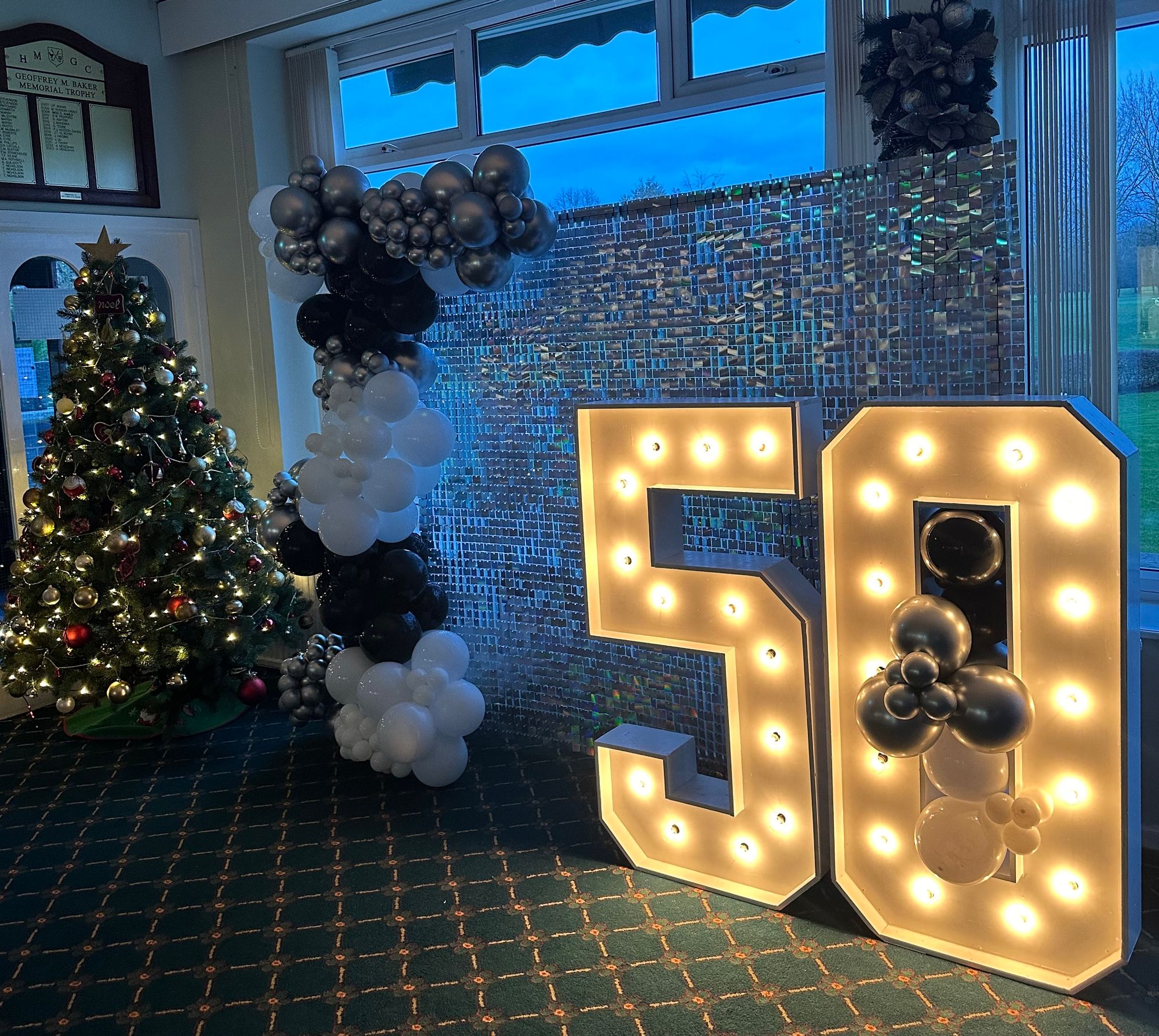 Indoor celebration setup with a decorated Christmas tree, black and white balloon arch, and illuminated number 50.